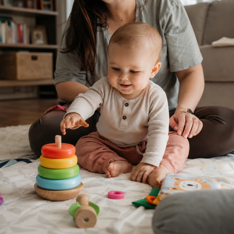 Baby playing with colorful wooden toys on a blanket, with an adult sitting behind