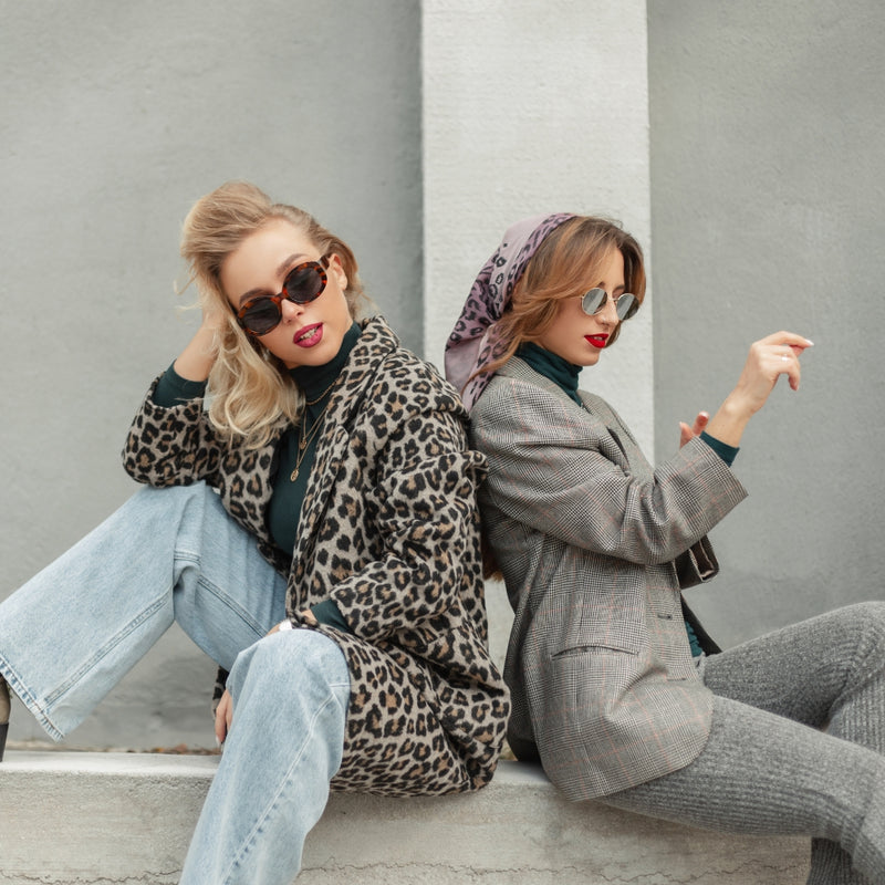 Two women posing against a gray wall, one in a leopard print coat and sunglasses, the other in a gray suit and headscarf.