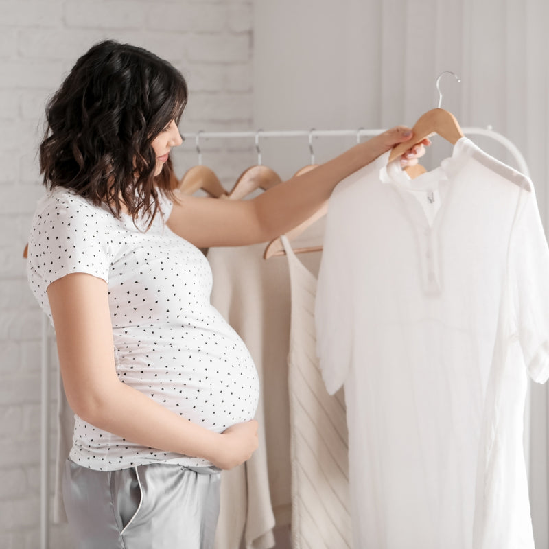 Pregnant woman selecting a white shirt from a rack in a room.