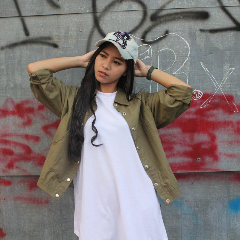 Person wearing a green jacket and white dress with a cap, standing against a graffiti-covered wall.